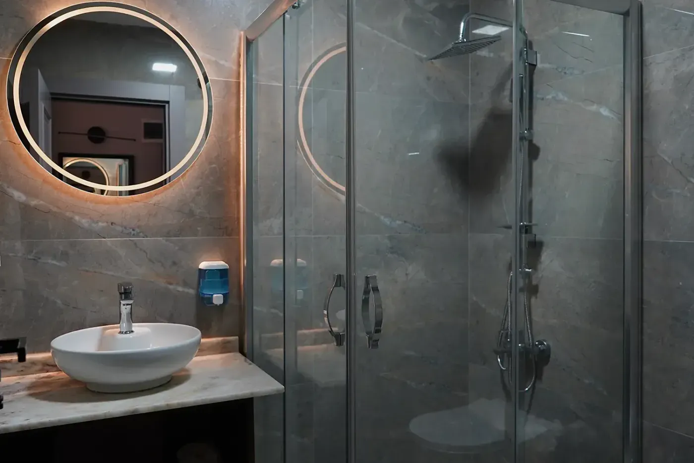 Deluxe Twin Oda banyosu: ışıklı yuvarlak ayna, mermer tezgâhta çanak lavabo ve cam duşakabin