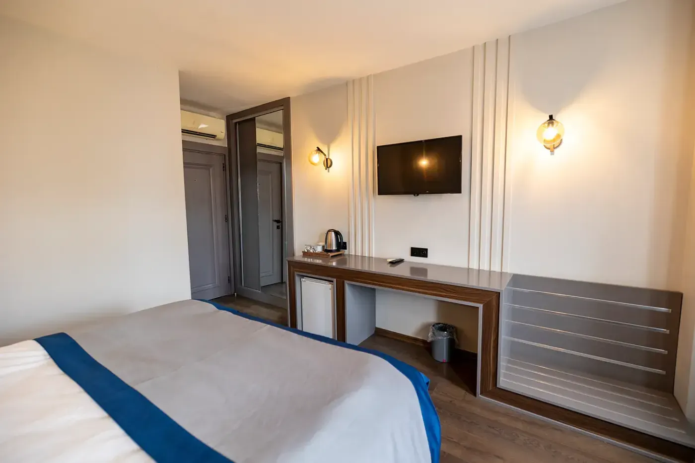 Deluxe Oda’nın giriş tarafı: aynalı gardırop, çalışma konsolu, mini buzdolabı ve duvara monte televizyon