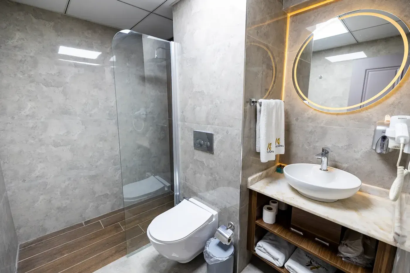 Deluxe Oda banyosu: cam bölmeli duş, asma klozet, mermer tezgâhta çanak lavabo ve ışıklı yuvarlak ayna