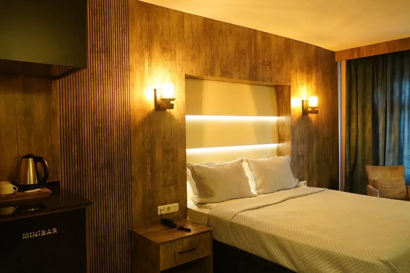 Junior Suit’in başucu: ışıklı deri başlık, iki aplik, ahşap komodin ve yanında su ısıtıcılı minibar ünitesi